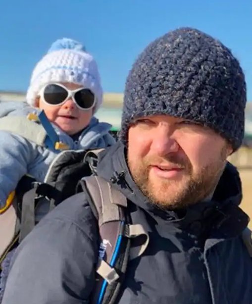 Alex bundled up in winter clothes with his baby on his back, both wearing woolly hats and sunglasses on a sunny day out.