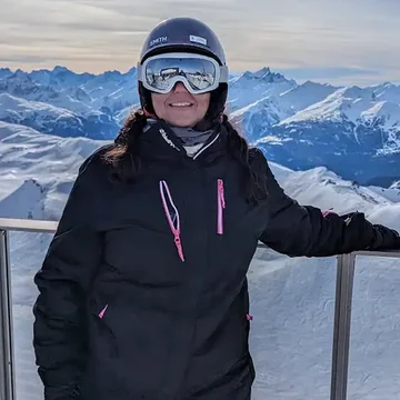 Lalita in full ski gear standing on a snowy mountain lookout, smiling with alpine peaks and valleys in the background.