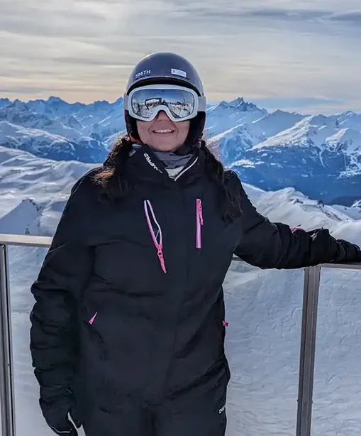 Lalita in full ski gear standing on a snowy mountain lookout, smiling with alpine peaks and valleys in the background.