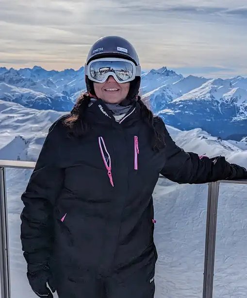 Lalita in full ski gear standing on a snowy mountain lookout, smiling with alpine peaks and valleys in the background.