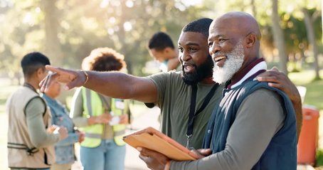 Two men collaborating during a community volunteer event, smiling as they discuss something on a tablet. One gestures ahead while the other looks on, highlighting how digital tools are supporting their teamwork and engagement.