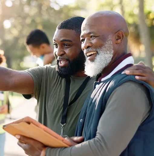 Two men collaborating during a community volunteer event, smiling as they discuss something on a tablet. One gestures ahead while the other looks on, highlighting how digital tools are supporting their teamwork and engagement.
