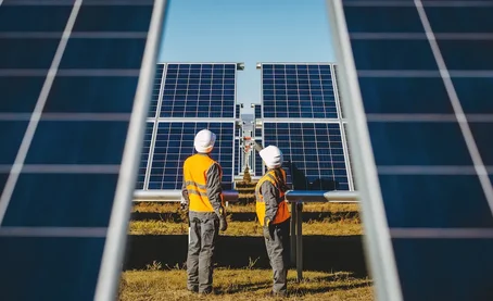 Two engineers in safety gear inspecting large solar panels at a solar farm on a clear, sunny day.