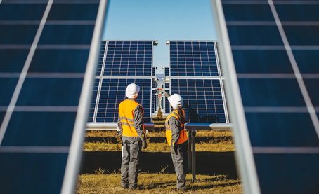 Two engineers in safety gear inspecting large solar panels at a solar farm on a clear, sunny day.