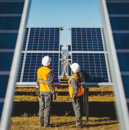 Two engineers in safety gear inspecting large solar panels at a solar farm on a clear, sunny day.