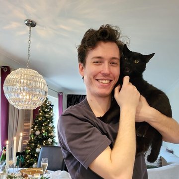 Will smiling and holding his black cat Anakin in a warmly lit dining room decorated for Christmas, with a set dinner table, champagne glasses, and a lit Christmas tree in the background.
