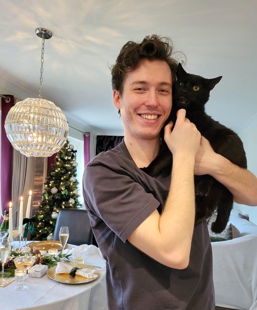 Will smiling and holding his black cat Anakin in a warmly lit dining room decorated for Christmas, with a set dinner table, champagne glasses, and a lit Christmas tree in the background.