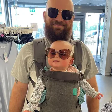 Rob Tagg wearing sunglasses and smiling indoors with his baby in a front carrier—both of them stylishly sporting oversized sunglasses in a lighthearted matching moment.