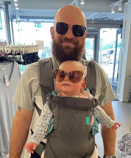 Rob Tagg wearing sunglasses and smiling indoors with his baby in a front carrier—both of them stylishly sporting oversized sunglasses in a lighthearted matching moment.