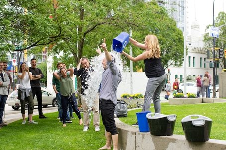 Woman pours ice water over man in an ice bucket challenge, while a group watches and laughs in a city park.
