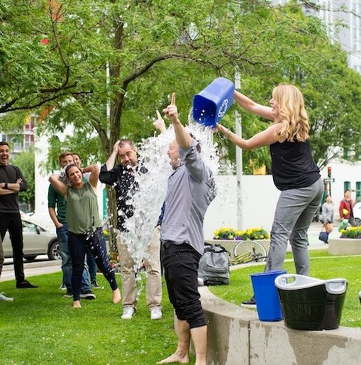 Woman pours ice water over man in an ice bucket challenge, while a group watches and laughs in a city park.