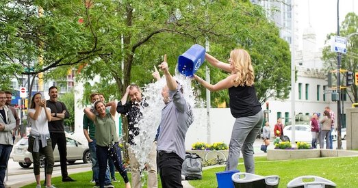 Woman pours ice water over man in an ice bucket challenge, while a group watches and laughs in a city park.