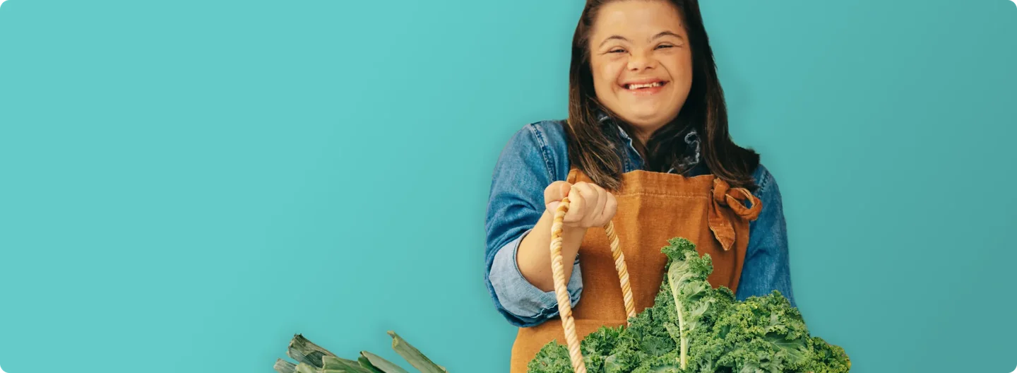 A smiling young woman wearing an apron holds a basket of fresh leafy greens and leeks, standing in front of a market stall filled with fruit and vegetables.