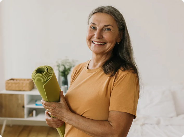 Smiling older woman holding a rolled-up yoga mat in a bright, peaceful room, suggesting wellness and an active lifestyle.