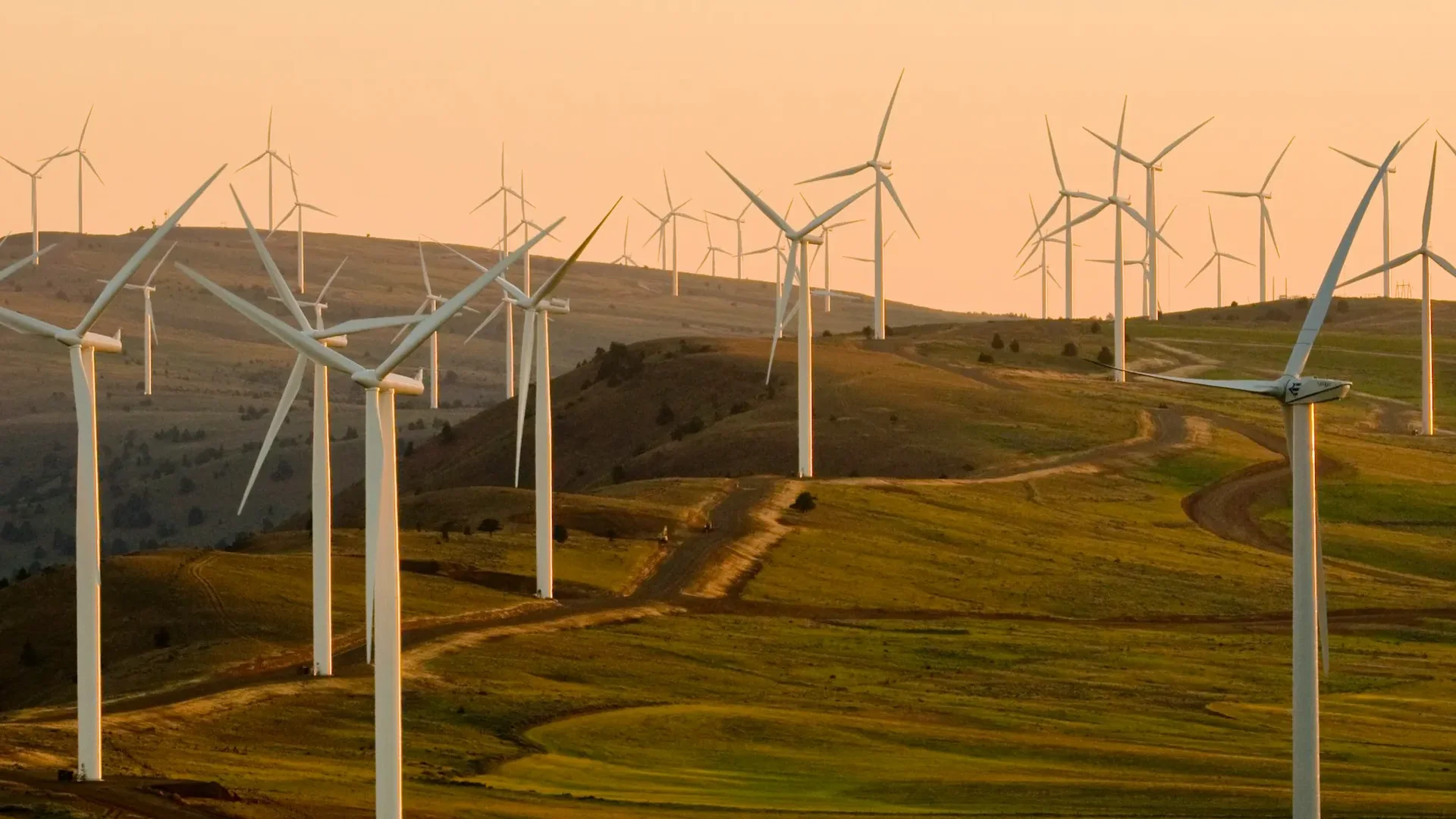 Wind turbines spread across rolling green hills at sunset, representing sustainable energy production, with the Hetzner logo and the words 'Server · Cloud · Hosting' in the corner.