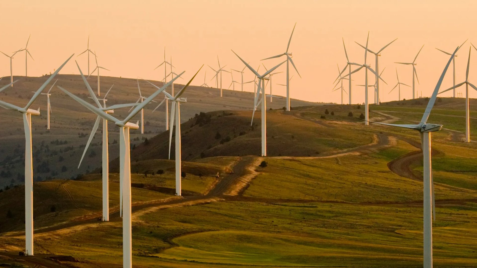 Wind turbines spread across rolling green hills at sunset, representing sustainable energy production, with the Hetzner logo and the words 'Server · Cloud · Hosting' in the corner.