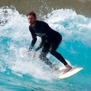 Gwilym riding a wave on a surfboard, captured mid-turn in a wetsuit surrounded by bright blue water and spray.