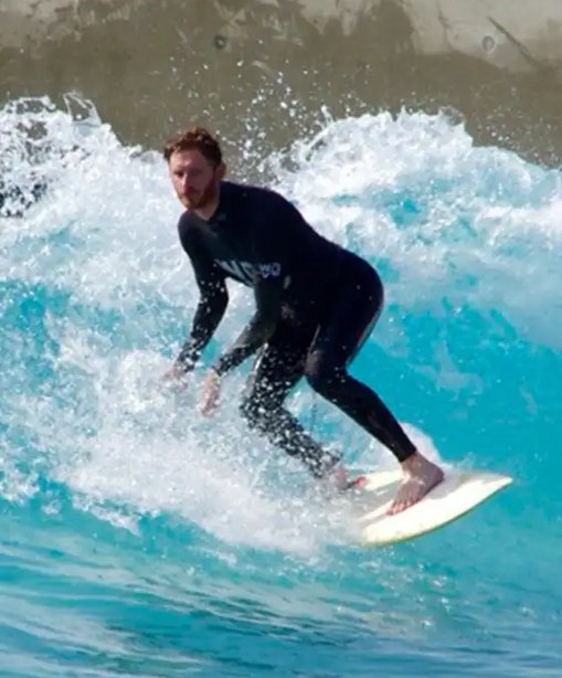 Gwilym riding a wave on a surfboard, captured mid-turn in a wetsuit surrounded by bright blue water and spray.