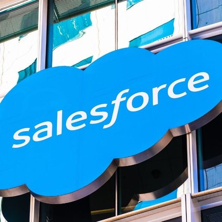 Close-up of a Salesforce logo on a blue cloud-shaped sign outside a modern glass office building.