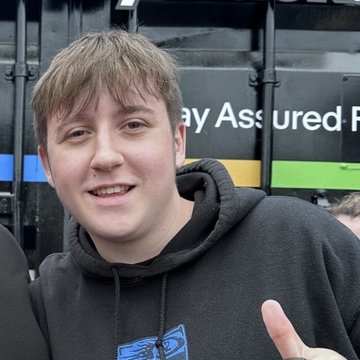 Toby is smiling and giving a thumbs-up while wearing a black hoodie, standing outdoors in front of a colourful background with text.