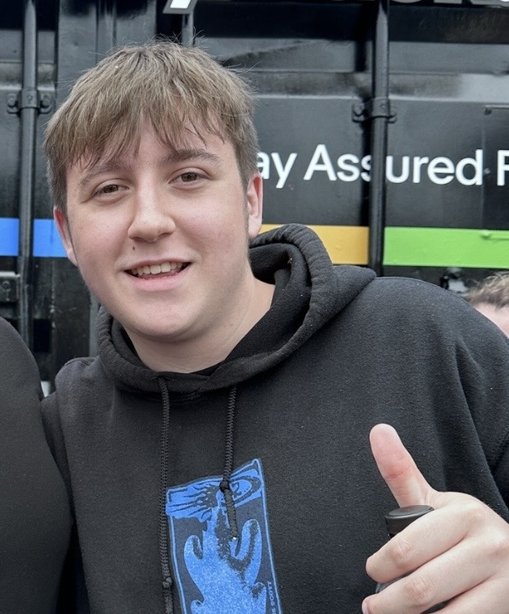 Toby is smiling and giving a thumbs-up while wearing a black hoodie, standing outdoors in front of a colourful background with text.
