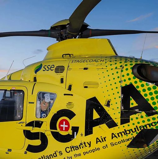 Close-up of a bright yellow Scotland’s Charity Air Ambulance helicopter with visible logos and markings, captured against a clear blue sky.