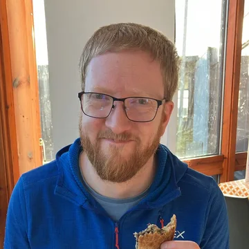 Peter, smiling and holding a half-eaten haggis toastie, sits at a table with an empty plate and cup during his honeymoon.