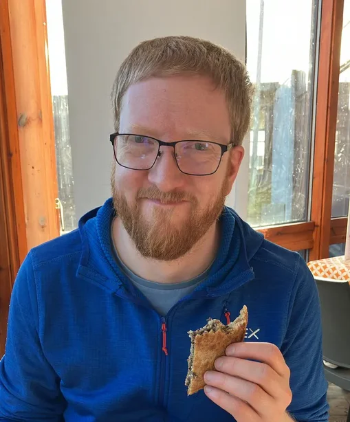 Peter, smiling and holding a half-eaten haggis toastie, sits at a table with an empty plate and cup during his honeymoon.
