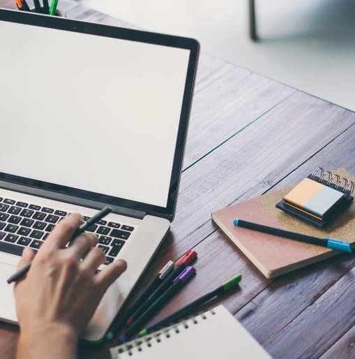 Person working at a wooden desk with a laptop, pens, and notepads, preparing to write or design on a blank screen.