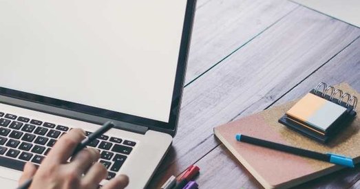 Person working at a wooden desk with a laptop, pens, and notepads, preparing to write or design on a blank screen.